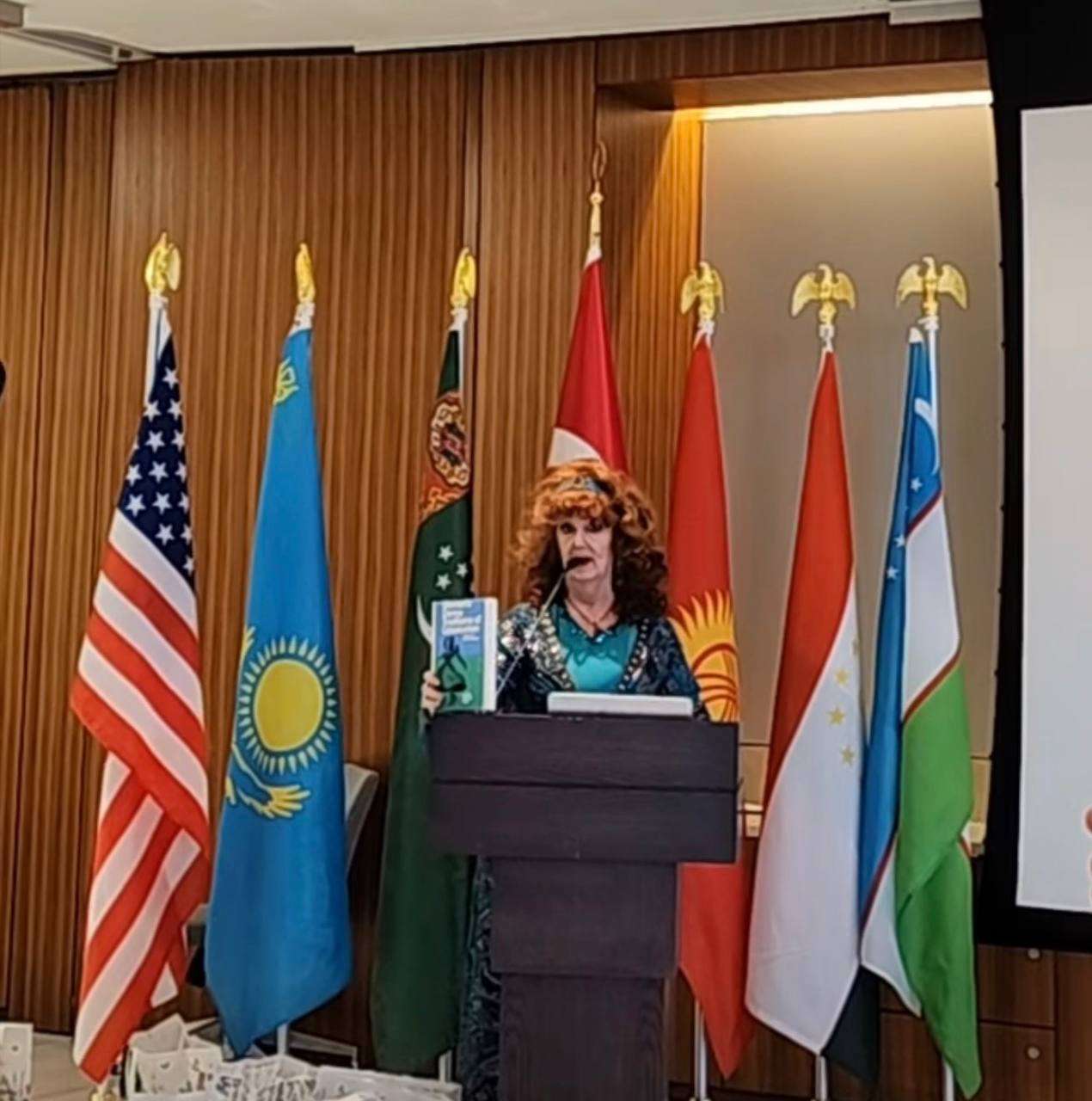 Speaker in traditional dress at a podium, framed by the flags of the United States and Central Asian nations