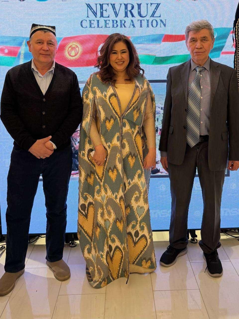 Three attendees before a Nevruz celebration screen, one wearing a traditional ikat gown