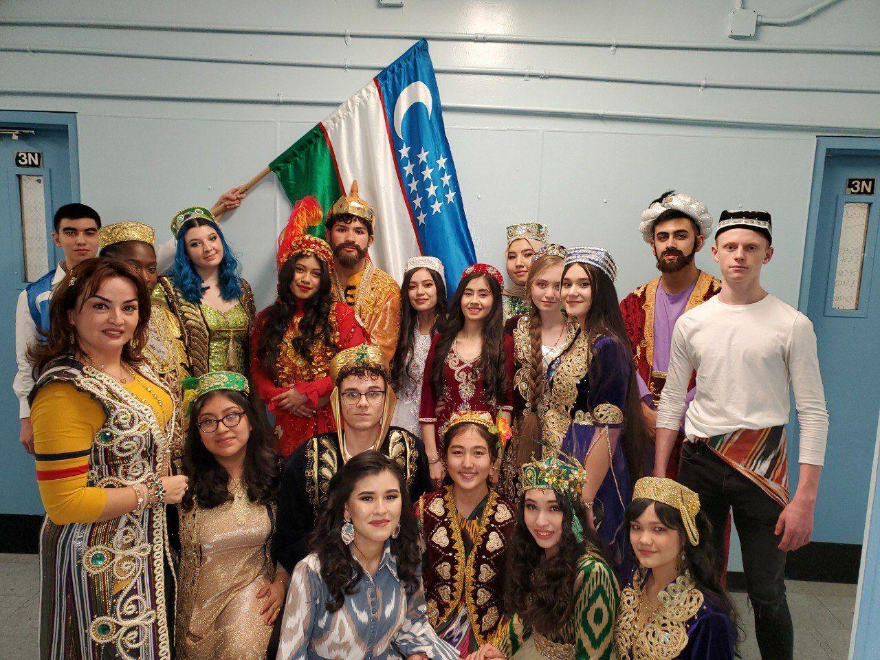 Young performers in historical costumes gathered before the flag of Uzbekistan in a school hallway